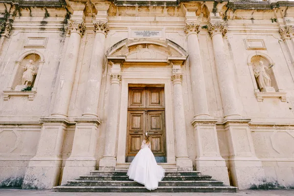 The bride in wreath standing on the stairs near the wooden door of the ancient temple in Prcanj, back view — Stock Photo, Image