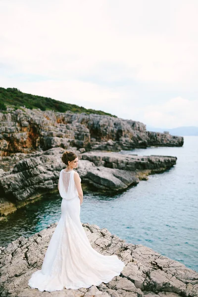 Uzun dantelli beyaz elbiseli düşünceli gelin denizin üzerindeki taş bir sahilde duruyor. — Stok fotoğraf
