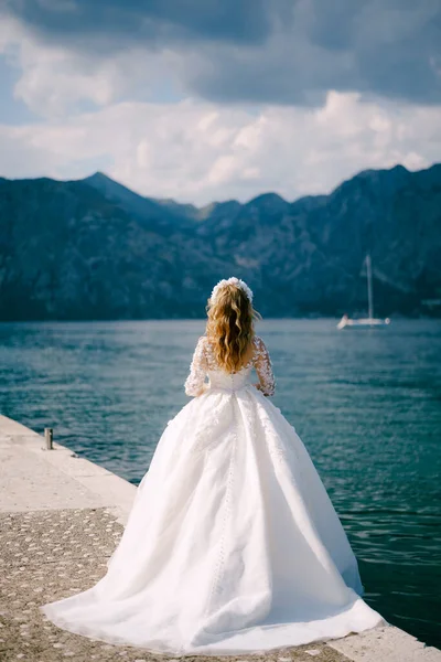 An elegant bride in wreath stands on a pier in the Bay of Kotor near the blue water, back view — Stock Photo, Image