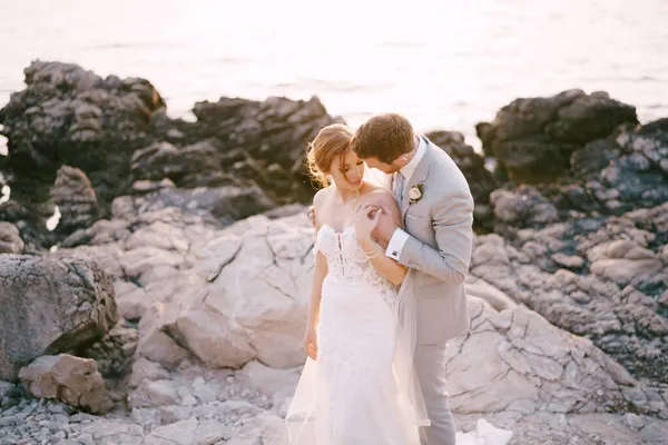 A vőlegény a menyasszony fölé hajolt, vállát a sziklás tengerparton ölelgette. — Stock Fotó