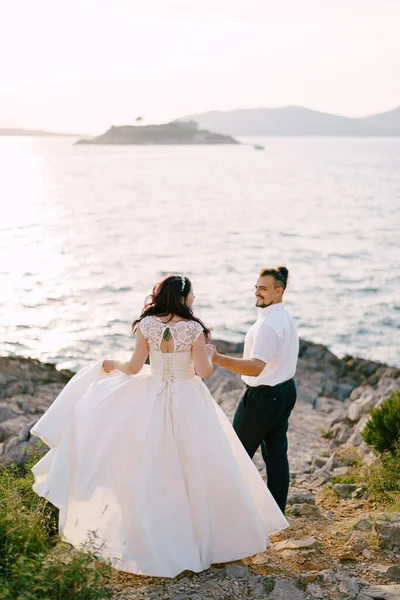 Жених и невеста идут держась за руки вдоль скалистого побережья к морю — стоковое фото
