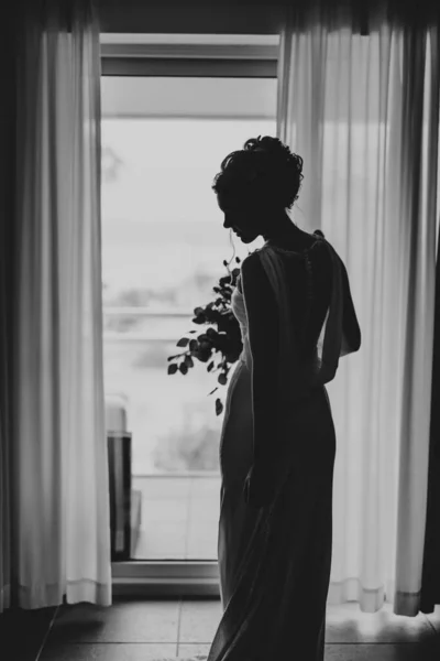 Krásná nevěsta v šatech s kyticí květin stojí, otáčí se a dívá se na podlahu v ložnici na pozadí okna. Černobílá fotografie — Stock fotografie