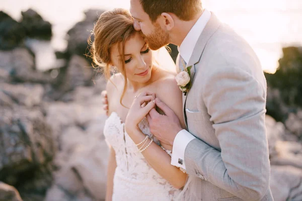 Жених целует невесту, обнимает ее за плечи на скалистом берегу моря — стоковое фото