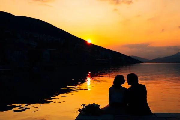 Silhouetten van koppel zitten op de pier boven het water tegen de achtergrond van bergen en zonsondergang — Stockfoto
