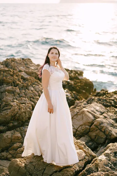 하얀 레이스 드레스를 입은 신부 가 바닷가 바위가 많은 해변에 서 있다 — 스톡 사진