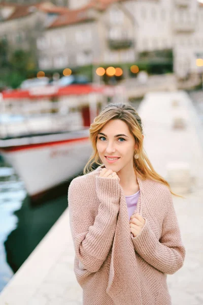 Joven mujer sonriente envuelta en una chaqueta caliente mientras está de pie en el muelle sobre el fondo de barcos y edificios — Foto de Stock