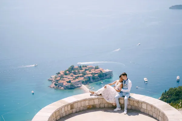 A noiva eo noivo estão sentados abraçando no deck de observação com vista para a ilha de Sveti Stefan — Fotografia de Stock