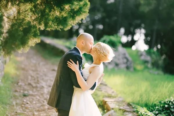Жених в черном костюме обнимает и целует невесту в белом платье, стоя на дорожке в парке на фоне зеленых деревьев — стоковое фото