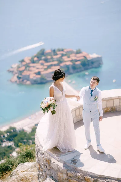 La mariée se tient sur le parapet de la plate-forme d'observation surplombant l'île de Sveti Stefan, le marié tient sa main — Photo