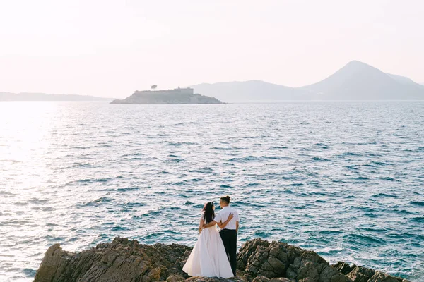 La novia y el novio se abrazan en las rocas y se miran entre sí. Vista trasera — Foto de Stock