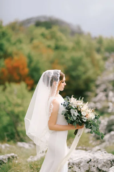 Pengantin dalam gaun putih dengan kerudung memegang karangan bunga di tangannya dengan latar belakang hijau dan pegunungan — Stok Foto