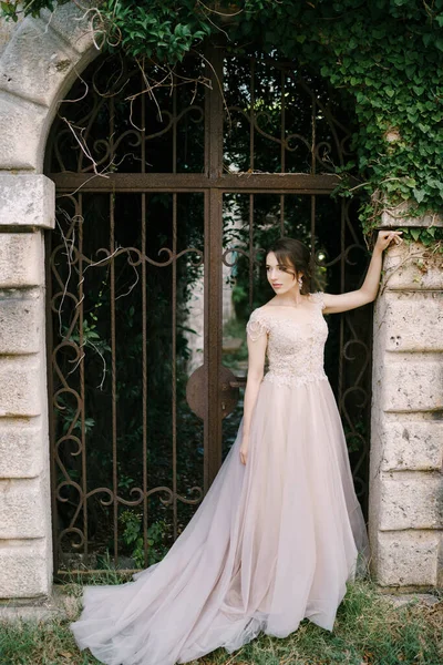 Noiva em um vestido de renda fica perto do portão forjado entrelaçado com hera — Fotografia de Stock