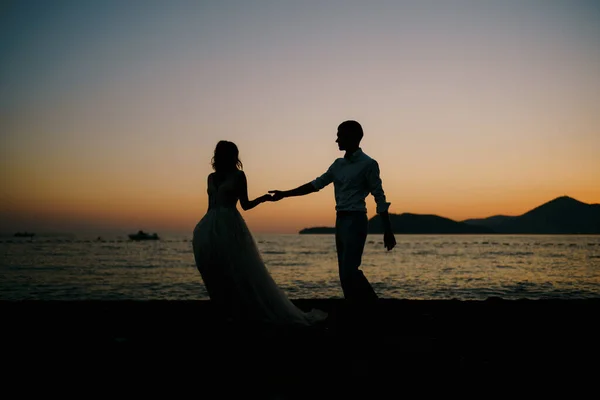 Silhouettes des mariés se tenant la main le long de la plage au coucher du soleil — Photo
