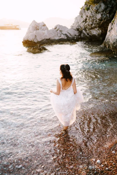 Noiva caminha no mar, segurando a bainha de seu vestido. Visão traseira — Fotografia de Stock