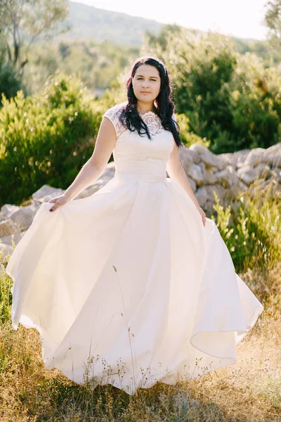 Bruden håller golven i en vit klänning mot en bakgrund av buskar och stenar — Stockfoto