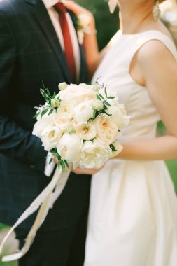 Mavi kareli takım elbiseli damat bir buket çiçekli beyaz elbiseli geline sarılıyor. Yakın plan.