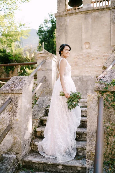 The bride with bridal bouquet standing on the stairs of the ancient bell tower near the church in Prcanj — Stock Photo, Image