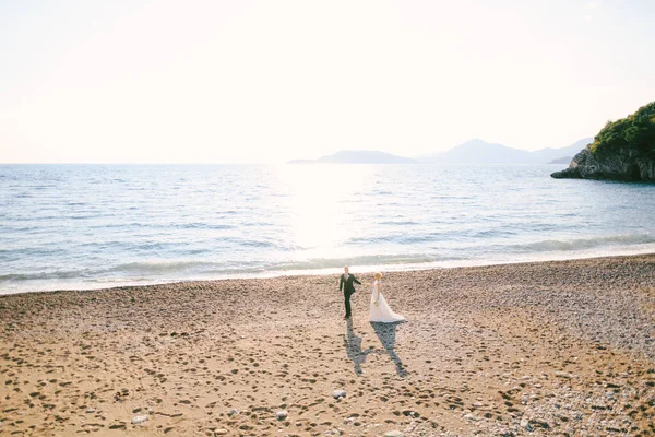 Bruidegom en bruid in een witte jurk met een boeket bloemen hand in hand lopen langs het strand aan zee tegen de achtergrond van groene bergen — Stockfoto