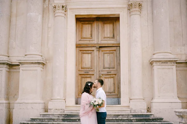 Жених целует невесту в лоб на фоне фасада здания — стоковое фото