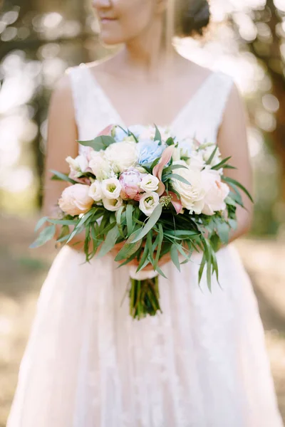 Seorang pengantin berdiri di sebuah kebun zaitun dan memegang di tangannya buket dengan mawar, peoni, lisianthus dan eucalyptus, close-up — Stok Foto