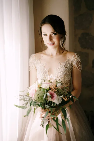 Novia en un hermoso vestido con un ramo de flores se encuentra cerca de la ventana. Retrato — Foto de Stock