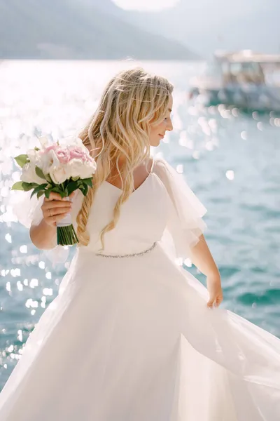 Μια νύφη με ένα ντελικάτο φόρεμα με ένα μπουκέτο στο χέρι στέκεται σε μια προβλήτα στον κόλπο του Κότορ — Φωτογραφία Αρχείου
