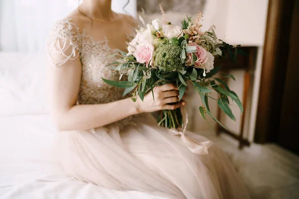 Noiva em um vestido de renda com um belo buquê de flores se senta na cama — Fotografia de Stock