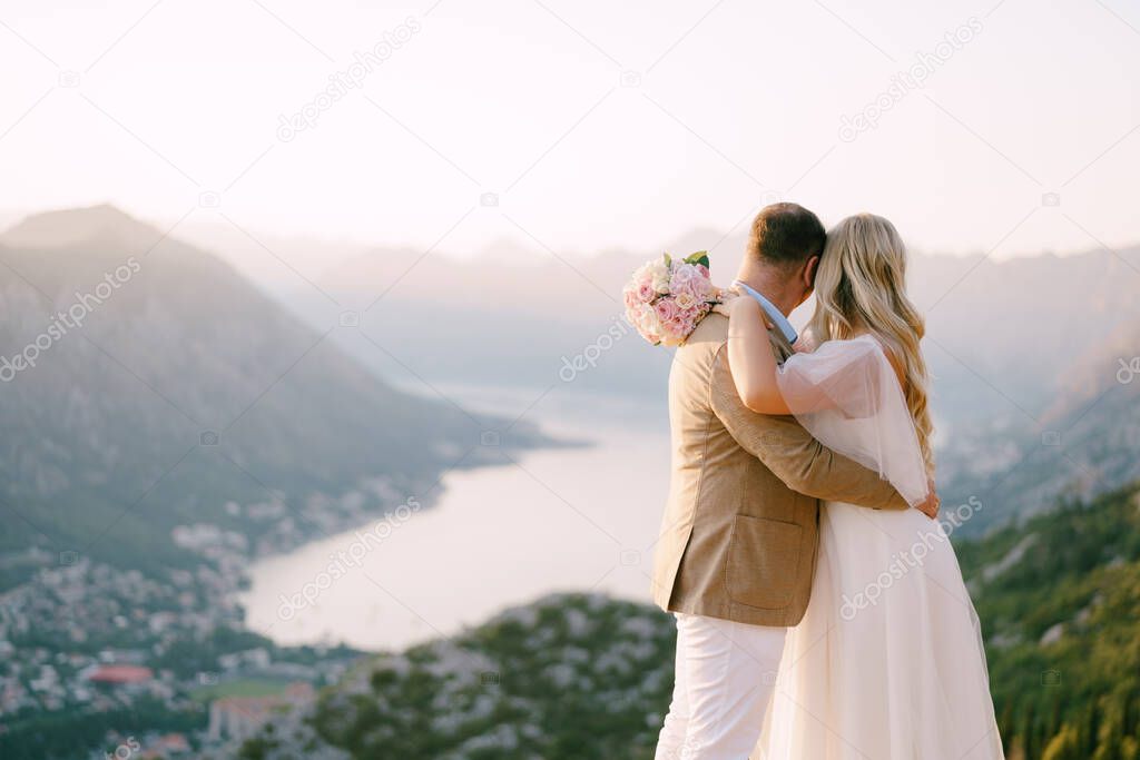 Bride and groom are hugging each other on the mountain and look at the bay