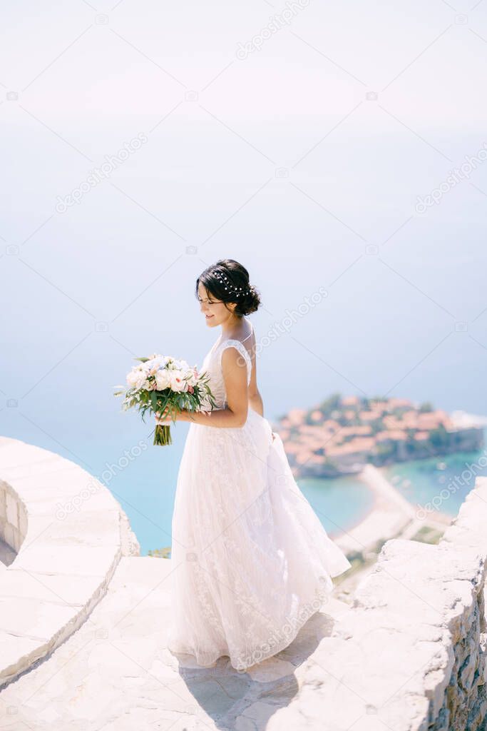 The bride with bridal bouquet stands on the observation deck overlooking the island of Sveti Stefan, back view