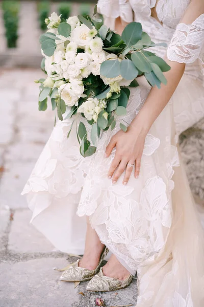 Pengantin dalam gaun renda dengan karangan bunga duduk di bangku batu. Close-up — Stok Foto