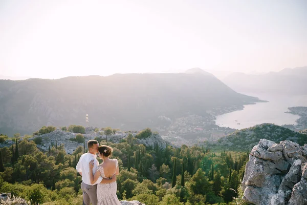 Sposo abbraccia la sposa su un'alta montagna sullo sfondo di un panorama della baia e degli alberi — Foto Stock