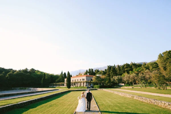 Novia y novio en un vestido blanco con un ramo de flores están caminando por el camino en un hermoso jardín con el telón de fondo de una antigua villa. Vista trasera —  Fotos de Stock