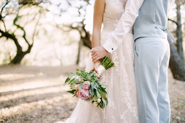 Gli sposi stanno abbracciando tra gli alberi, la sposa tiene bouquet da sposa, primo piano — Foto Stock