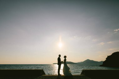 Damat, gelinin önünde elinde bir buket çiçekle gün batımında deniz kenarındaki setin kenarındaki dağın arka planında duruyor.