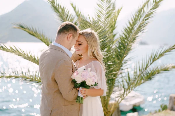 Le marié et la mariée souriante avec un support de bouquet sur la jetée à l'arrière-plan d'un palmier et câlin tendrement — Photo