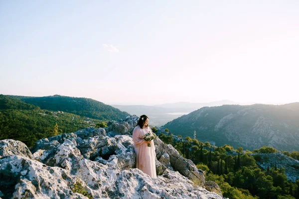 Mariée avec un bouquet se tient sur les pierres et regarde les montagnes — Photo