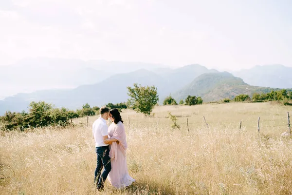 Жених обнимает невесту в поле на фоне гор — стоковое фото