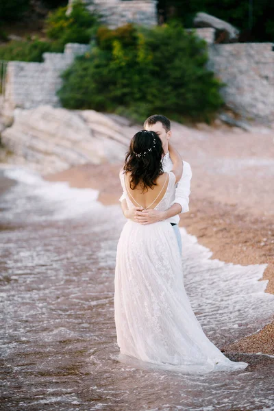 Groom abraça a noiva enquanto está de pé na praia perto da água — Fotografia de Stock