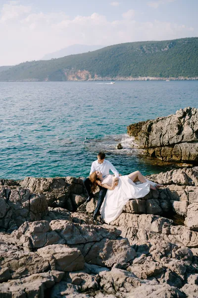 Bruden ligger på knä brudgummen sitter på stenarna vid havet — Stockfoto