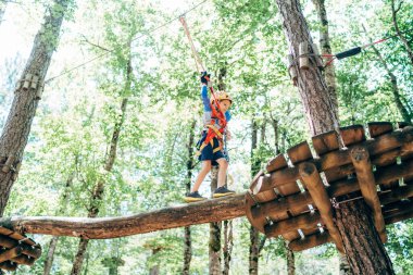 Çocuk iplere tutunarak havadan ahşap bir platforma doğru yürüyor.