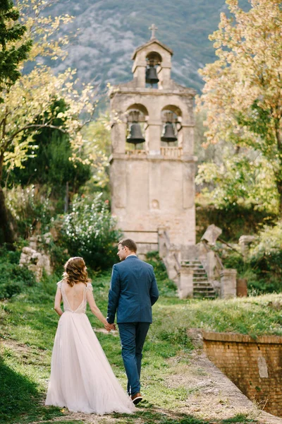 Bride and groom walk through the park to the old chapel. Back view