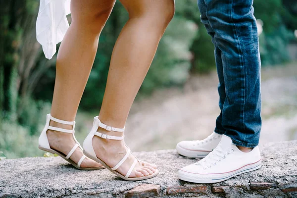 Kvinnliga fötter i sandaler bredvid manliga fötter i sneakers står på en sten gräns i parken — Stockfoto