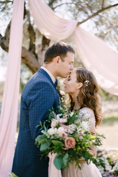 Groom embrasse la mariée sur le front avec un bouquet de fleurs sous un arbre. Portrait — Photo