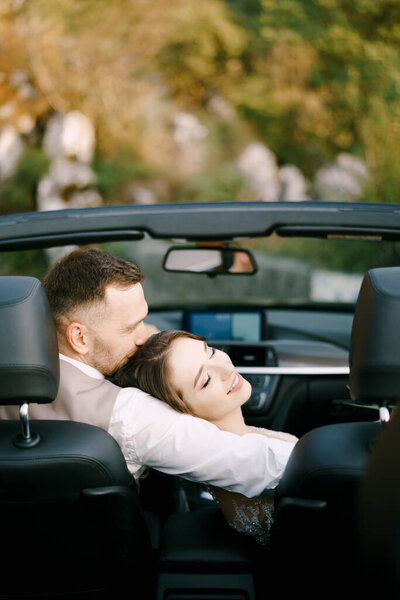 Groom hugs bride who has laid her head on his shoulder in a convertible