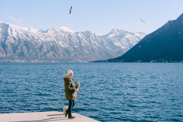 Girl in a jacket stands on the pier and feeds the seagulls