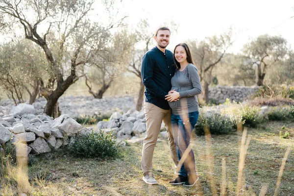 Sorridente donna incinta in piedi in un abbraccio con un uomo su uno sfondo di alberi — Foto Stock