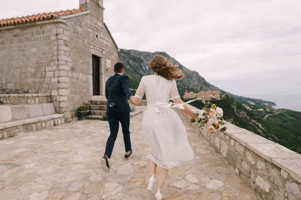 Mariée et fiancée avec un bouquet de fleurs courent à l'église sur la montagne. Vue arrière — Photo