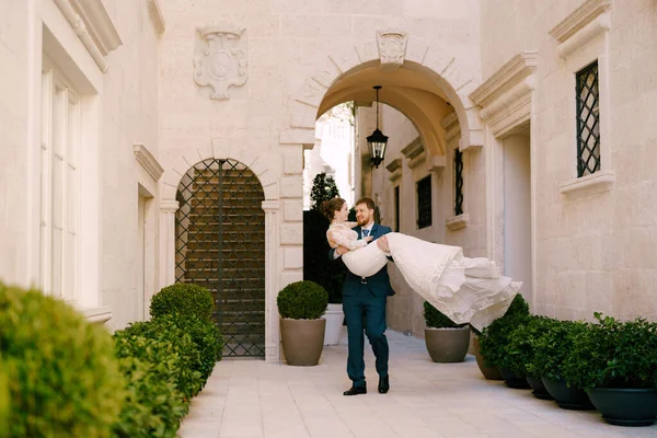 Ženich nosí nevěstu v náručí na nádvoří domu s oblouky — Stock fotografie