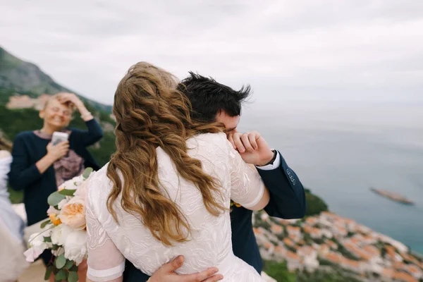 Bräutigam umarmt Braut mit einem Blumenstrauß, während sie auf dem Berg steht. Rückseite — Stockfoto