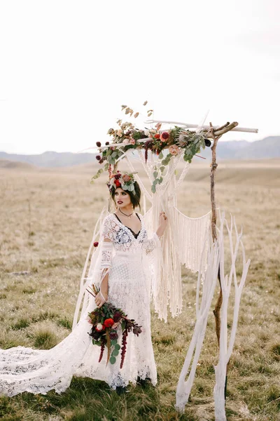 Mempelai dalam gaun wicker putih dan karangan bunga berdiri di lapangan di samping lengkungan pernikahan — Stok Foto